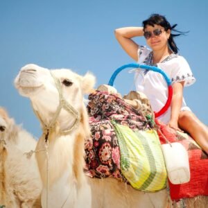 Young female tourist in tunic and sunglasses poses with camels against background Mediterranean sea